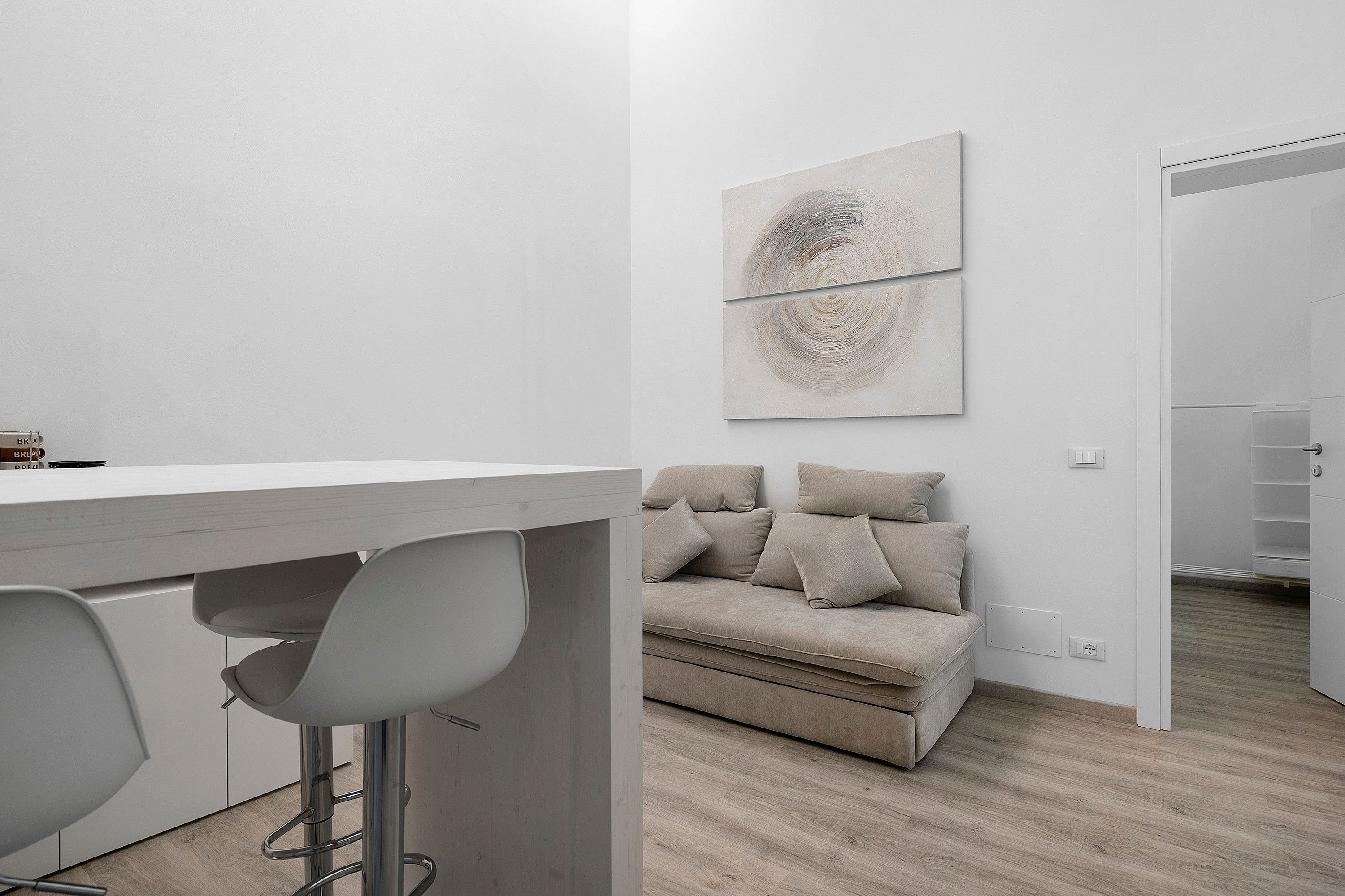 A modern, minimalist room with a white desk, two bar stools, a beige sofa with cushions, and a wall-mounted abstract painting.