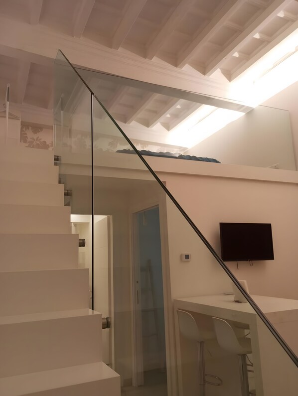 Eine moderne Treppe mit Glasgeländern, die zu einer zweiten Etage führt. An der Wand ist ein Flachbildfernseher angebracht.