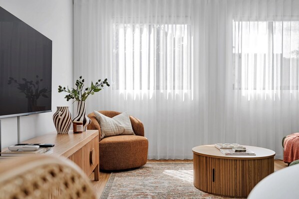 A modern living room with a flat-screen TV, a brown armchair, a round wooden coffee table, and a large window with sheer curtains. - Olive & Ochre | Warm Coastal Retreat (Shoalwater)