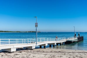A pier extending into the sea with a white railing, a sign warning of shallow water, and a clear blue sky. - Olive & Ochre | Warm Coastal Retreat (Shoalwater)