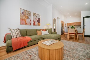 A living room with a green sofa, a round coffee table, and a dining area with wooden chairs. - Olive & Ochre | Warm Coastal Retreat (Shoalwater)