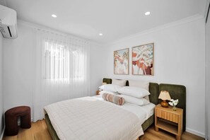 A bedroom with a bed, a nightstand, a lamp, a chair, and a window with curtains. - Olive & Ochre | Warm Coastal Retreat (Shoalwater)