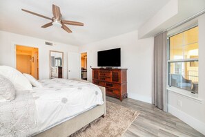 Ein Schlafzimmer mit einem Bett, einem Deckenventilator, einem Fernseher, einer Kommode und einem großen Fenster.