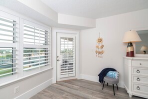 A room with a white door, a window with blinds, a lamp on a dresser, and a hanging decoration. - A301 Links at Harbour Village 3/3 condo. (Ponce Inlet)