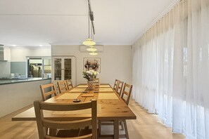 A dining area with a wooden table, chairs, a vase with flowers, and a kitchen in the background. - Beachside 4-Bed House With Pool In Warnbro (Warnbro)