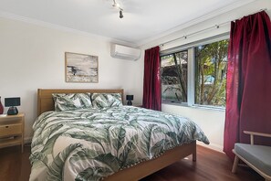 A bedroom with a bed, a nightstand, a lamp, a window with curtains, and a wall-mounted air conditioner. - Beachside 4-Bed House With Pool In Warnbro (Warnbro)