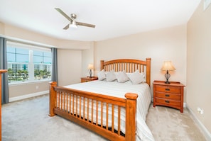 A bedroom with a wooden bed, a ceiling fan, two bedside tables with lamps, a large window with a view, and a carpeted floor. - A506 Links South in Ponce Inlet, FL (Ponce Inlet)