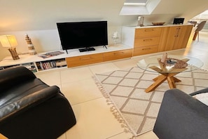 Ein modernes Wohnzimmer mit Flachbildfernseher, Ledersofas, einem Glastisch und einer Küchenzeile mit Holzschränken.