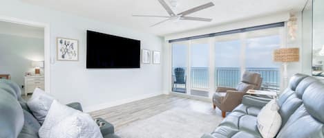 A spacious living room with a large flat-screen TV, a ceiling fan, and a balcony with a view of the ocean.