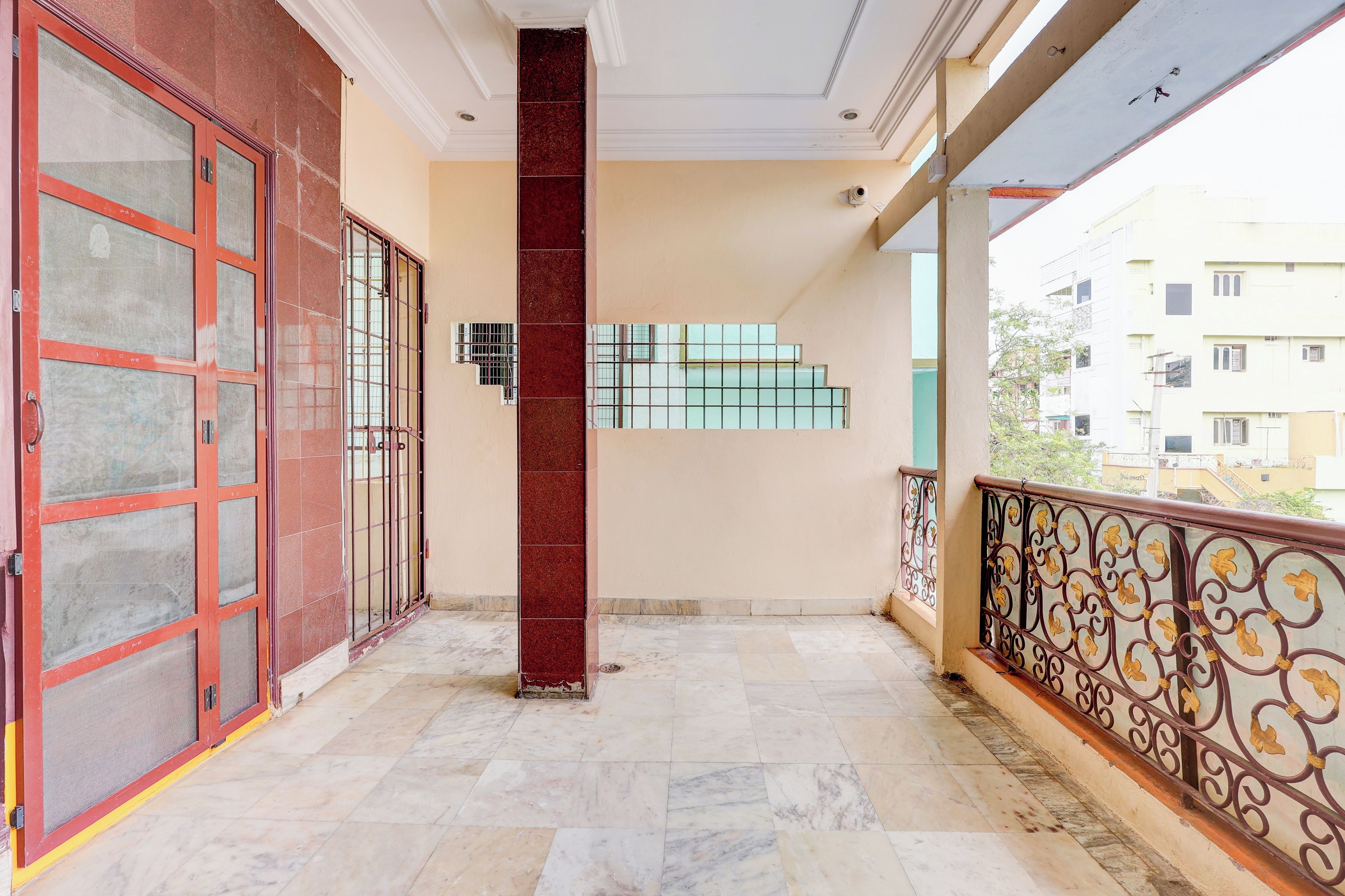 Un balcón con piso de baldosas, una puerta roja con reja metálica y vista a un edificio en el fondo.