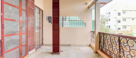 A balcony with a tiled floor, a red door with a metal grill, and a view of a building in the background.