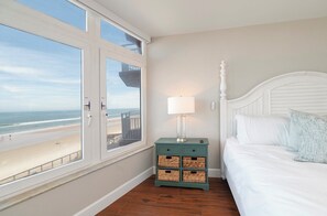 Een slaapkamer met een bed, een nachtkastje met een lamp, uitzicht op het strand en een raam met uitzicht op de oceaan.