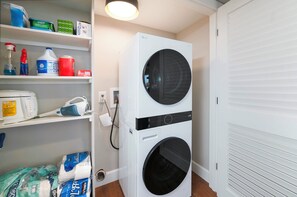 Un lavadero con dos lavadoras apiladas, un estante con productos de limpieza y una puerta blanca.