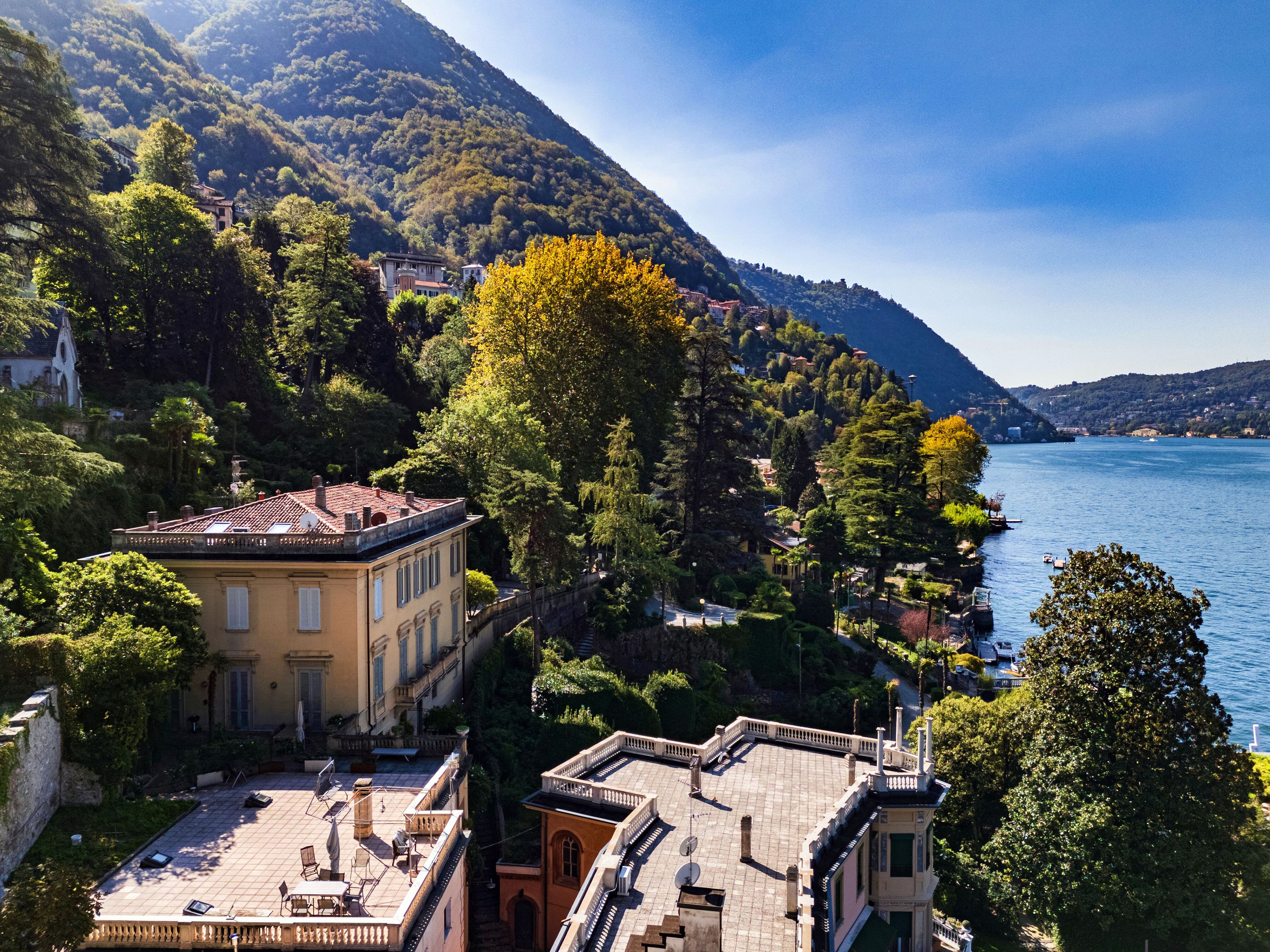 A coastal building with a terrace overlooking a body of water, surrounded by lush greenery and hills.