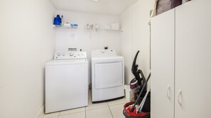 A laundry room with two white washing machines, a shelf with cleaning supplies, and a mop leaning against the wall. - Dimucci Twin Towers 1503 (Daytona Beach Shores)