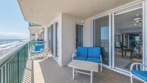 A balcony with a sofa, chairs, and a table, overlooking a beach and ocean. - Dimucci Twin Towers 1503 (Daytona Beach Shores)