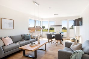 Ein modernes Wohnzimmer mit einer Couch, einem Couchtisch und Sesseln. Es gibt einen Essbereich mit Tisch und Stühlen. Große Fenster bieten einen Ausblick ins Freie.