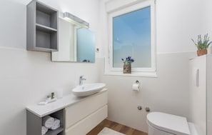 Ein modernes Badezimmer mit einem weißen Waschbecken, Waschtisch und einer Toilette. Ein Fenster bietet Blick auf den Himmel, auf dem Fensterbrett steht eine Pflanze.