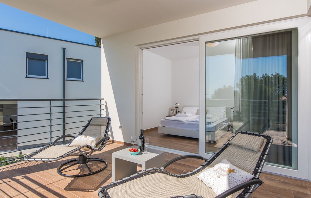 A balcony with two lounge chairs, a small table with a bowl of fruit and a glass of wine, and a bedroom with a bed and a sliding glass door leading to the balcony.