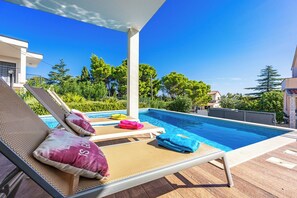 A poolside area with lounge chairs and towels, surrounded by greenery and buildings.