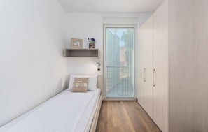 A narrow room with a bed, a small shelf with items, and a sliding door leading to an outdoor area.