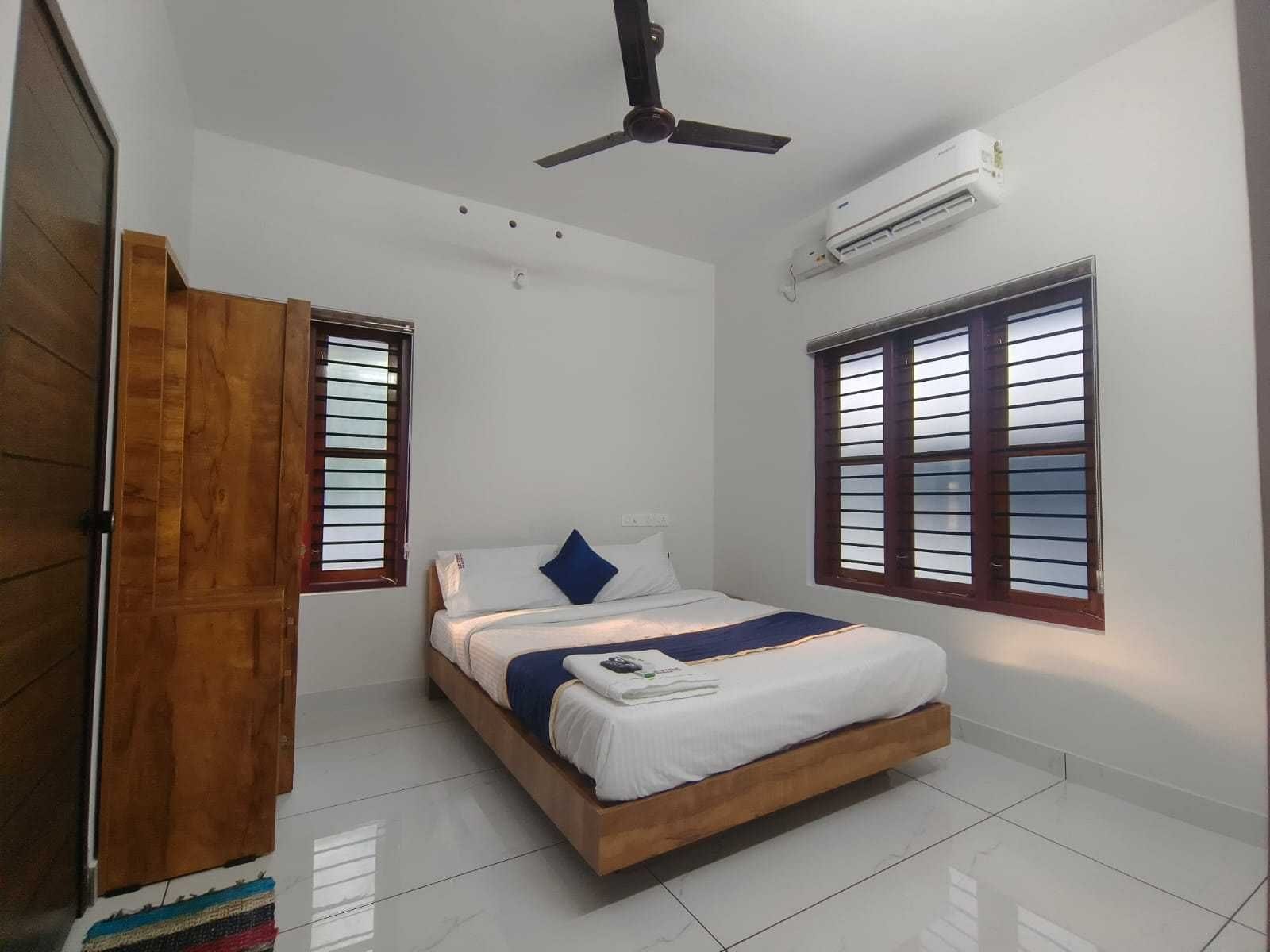 Habitación con cama, armario de madera, ventilador de techo, aire acondicionado y dos ventanas con persianas.