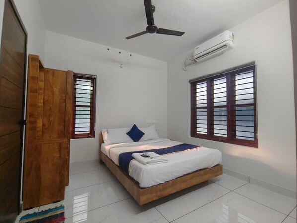 A bedroom with a bed, wooden wardrobe, ceiling fan, air conditioning unit, and two windows with blinds.