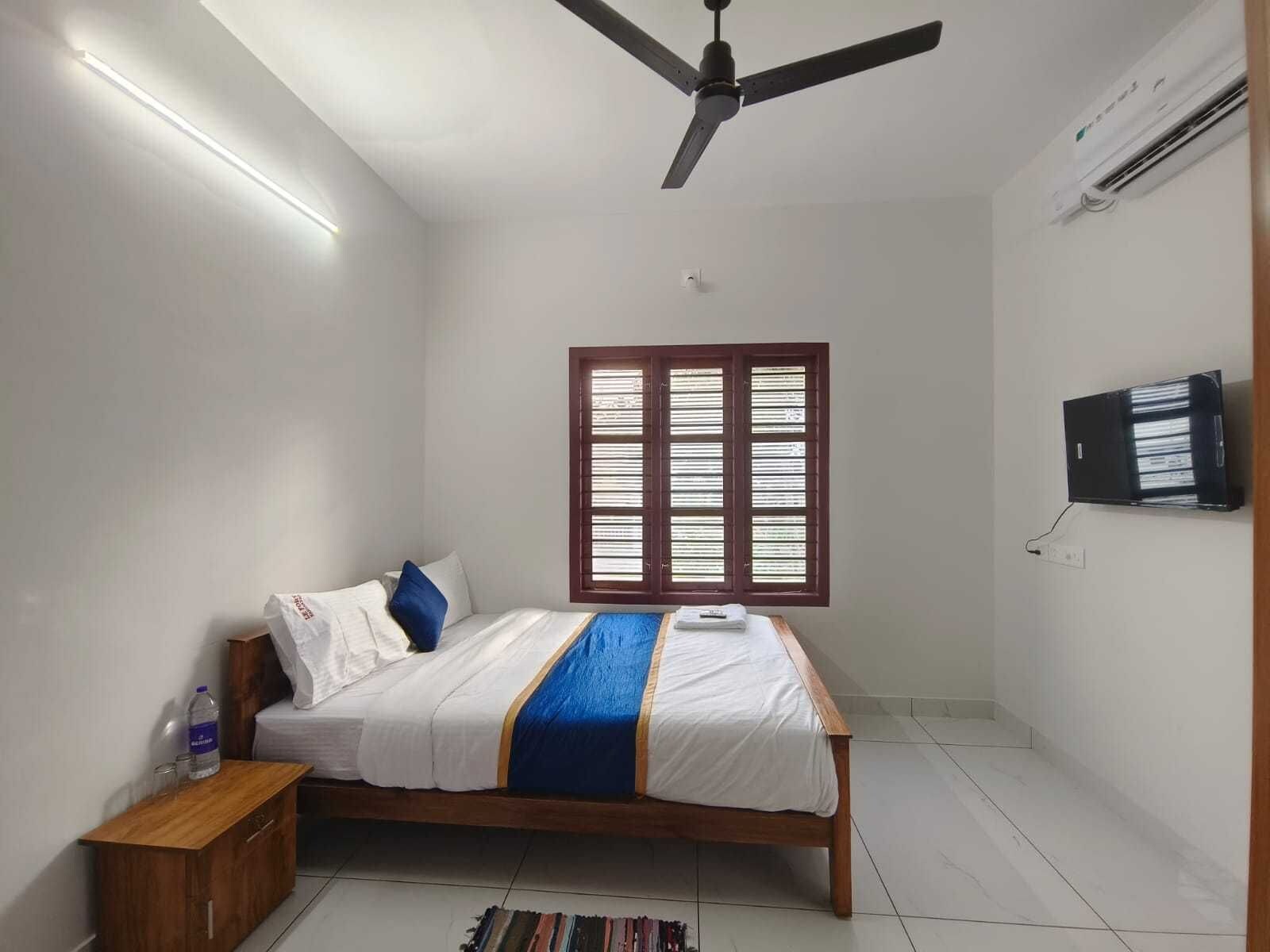 Ein Schlafzimmer mit einem Bett, einem Nachttisch, einem an der Wand montierten Fernseher, einem Deckenventilator und einem Fenster mit Jalousien.