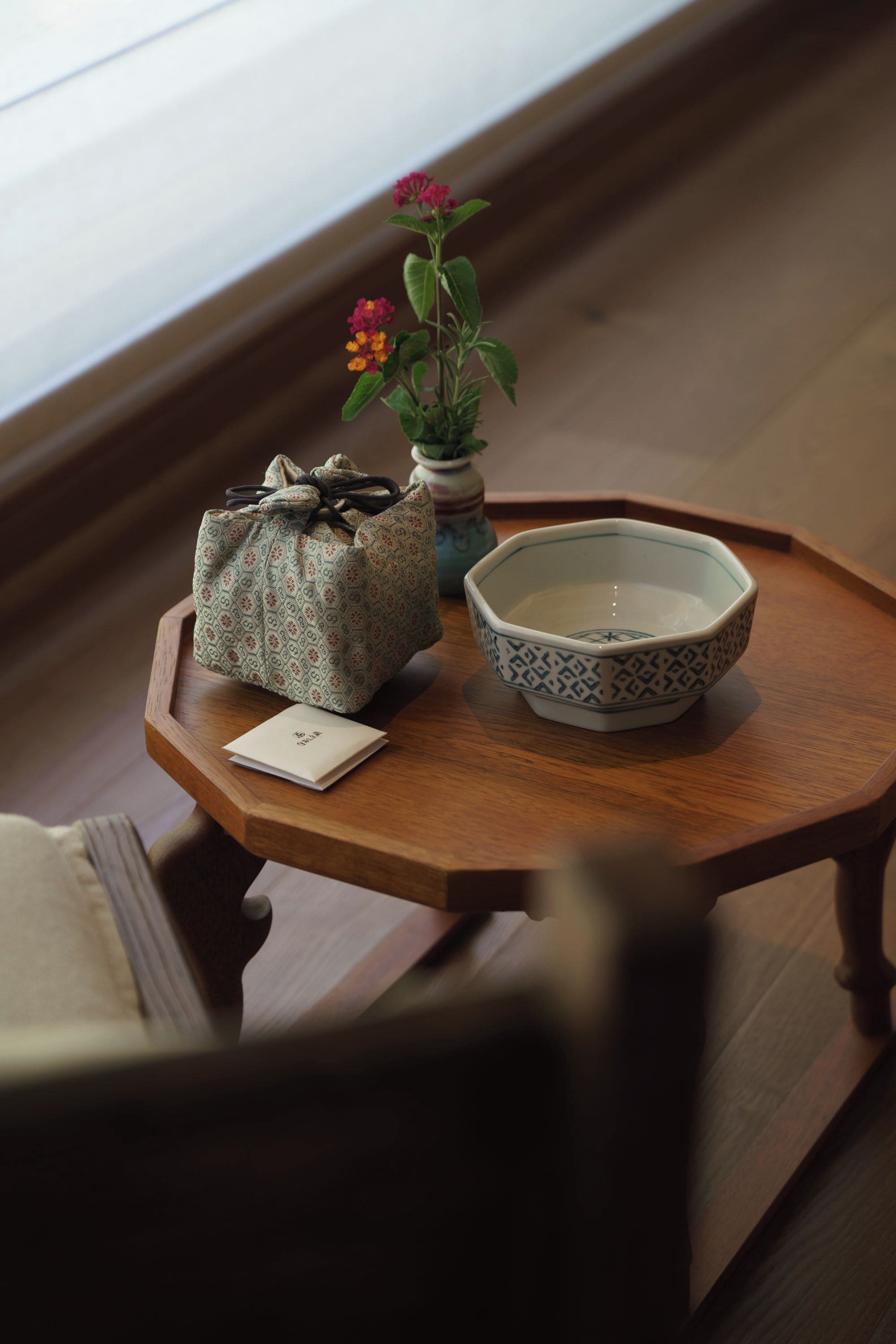 Una pequeña mesa de madera con un bolso estampado, un cuenco y un florero con flores.
