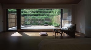 A room with a bed, a small round table, and a chair. There is a large sliding glass door leading to an outdoor area with a bamboo fence and greenery.