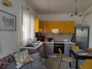 Cuina senzilla amb armaris grocs, un rentaplat, una placa de cocció i un frigorífic. Hi ha un menjador amb cadires i un sofà amb coixí estampat.