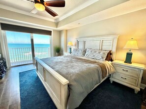 A bedroom with a large bed, a nightstand with a lamp, a view of the ocean through a sliding door, and a ceiling fan. - Sanibel Condo #1002 3/3 Direct Oceanfront NE Corner (Daytona Beach Shores)