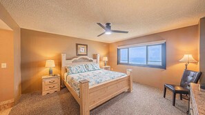 A bedroom with a wooden bed, two bedside tables with lamps, a ceiling fan, a window with a view, and a chair. - #1004 Seychelles 3/3 Oceanfront Traffic Free Beach (Daytona Beach Shores)