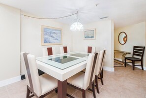 Un comedor con una mesa de vidrio, seis sillas, dos cuadros, una araña y un espejo en la pared.