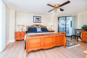 A bedroom with a wooden bed, two bedside tables with lamps, a ceiling fan, a framed picture on the wall, and a view through a glass door. - #101 Seychelles 2/2 Oceanfront Traffic Free Beach (Daytona Beach Shores)