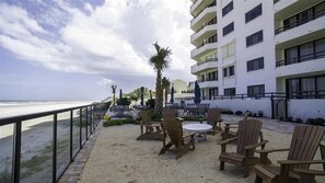 Eine Strandterrasse mit Sitzgelegenheiten im Freien, Blick auf das Meer und ein mehrstöckiges Gebäude im Hintergrund.
