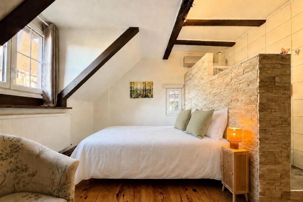 A bedroom with a bed, a nightstand, a lamp, a chair, a window, and a stone wall.