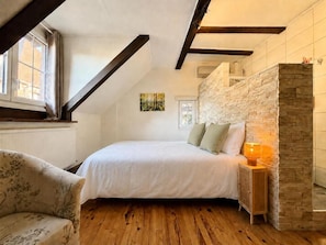 A bedroom with a bed, a nightstand, a lamp, a chair, a window, and a stone wall. - La Maison du Chat Perché  (INGERSHEIM)