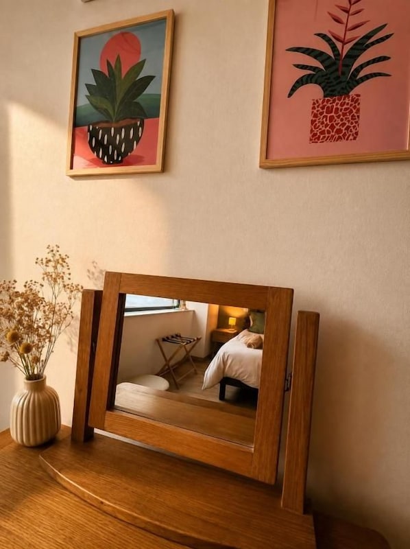 Un espejo en marco con el reflejo de una habitación, un jarrón con flores secas y dos cuadros en la pared.