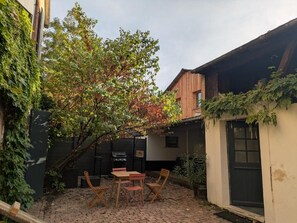 Un patio con piso de ladrillo, una mesa de madera con sillas, una parrilla y un árbol.