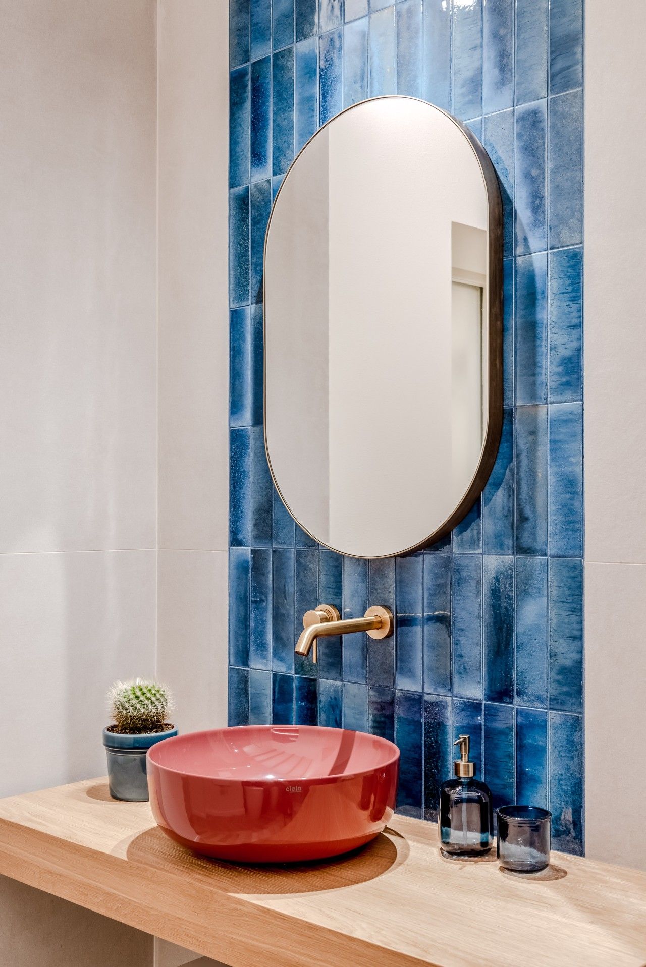 Ein Badezimmer mit rundem Spiegel, einem roten Waschbecken, einem kleinen Kaktus und einer Seifenflasche.