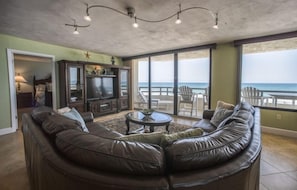 En stue med en buet lædersofa, et sofabord og et fjernsyn. Der er en balkon med udsigt over havet.