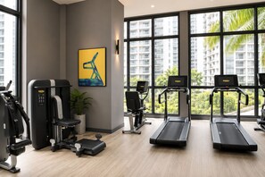 A gym with exercise equipment, a treadmill, and a large window with a view of buildings and trees. - Skyline Service Suite KLCC at The Mews (Kuala Lumpur)