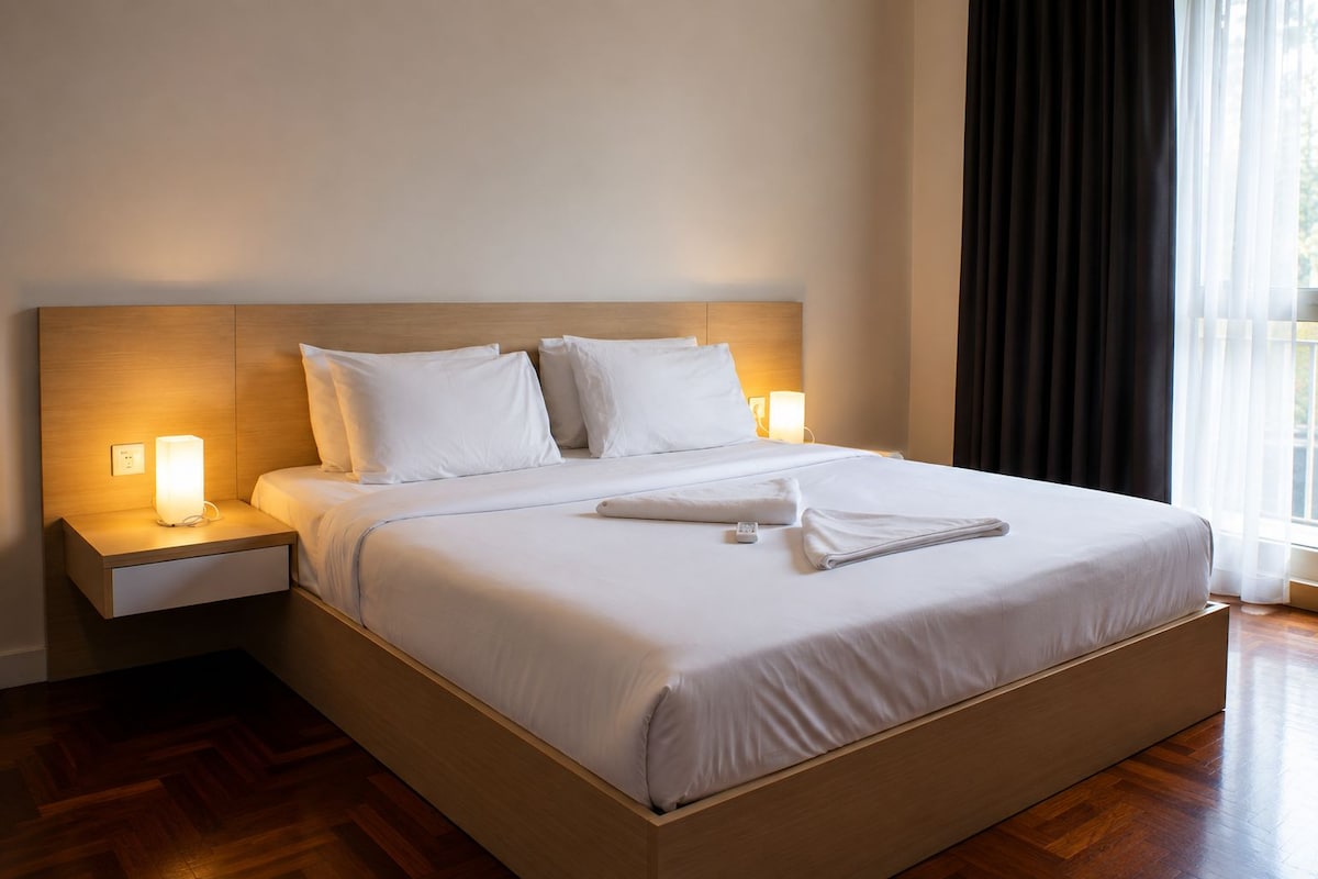 A hotel room with a large bed, two bedside tables with lamps, a wooden headboard, and a view of the outdoors through a window with curtains.
