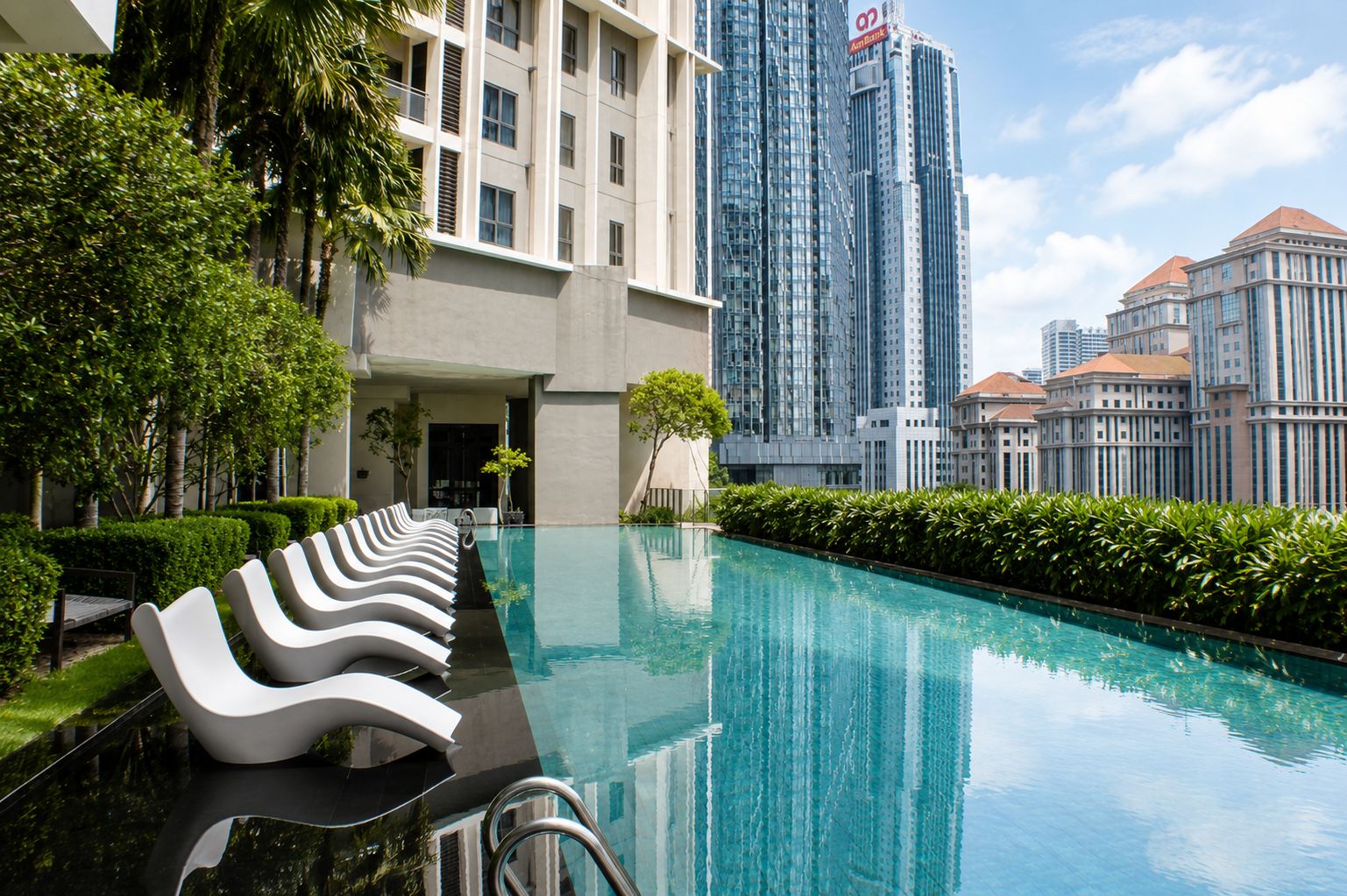 Una piscina con sillones de descanso modernos, rodeada de follaje y edificios altos al fondo.
