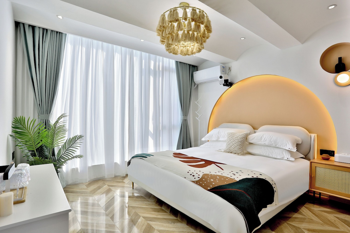 Habitació d'hotel moderna amb un llit gran, una taula de treball, una planta en test i una finestra gran amb cortines.