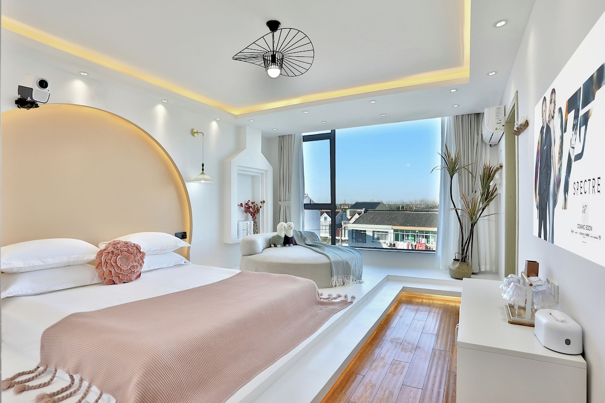 A modern hotel room with a large bed, a bedside table with amenities, a view of the city, and a unique ceiling light fixture.