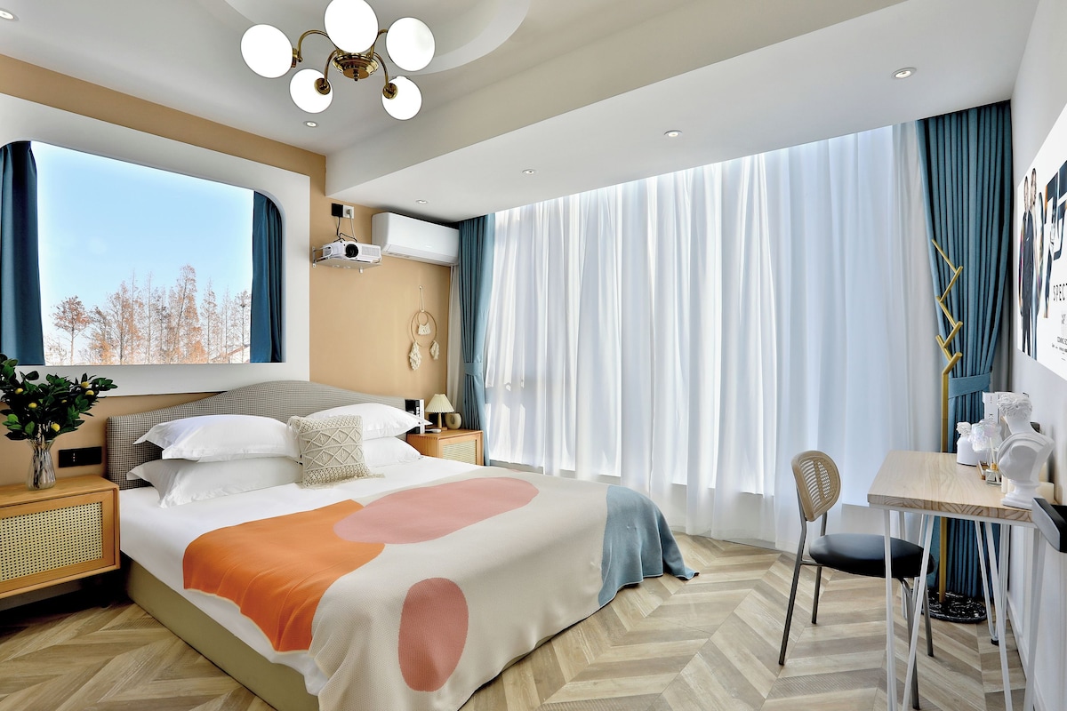 A modern hotel room with a large bed, a desk with a chair, a window with curtains, and a ceiling light fixture.