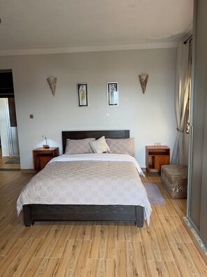 A bedroom with a large bed, wooden flooring, a nightstand, a bedside table, and a small wooden cabinet. - Lake view 2-bedroom apartment with power backup in vibrant Kampala neighborhood (Kampala)