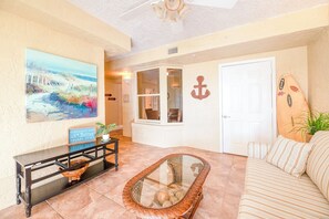A living room with a beach-themed painting, a glass coffee table, and a striped sofa. - Towers 4104 in Ponce Inlet, FL (Ponce Inlet)